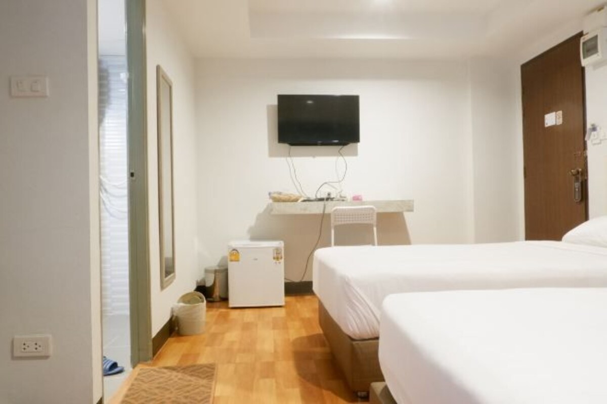 A hotel room with two beds, a TV mounted on the wall, a small desk with a chair, and a refrigerator.