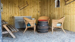 Et rustikt værelse med trægulv, to fletstole, en træfade, en træbænk og en lille, hvid hylde.