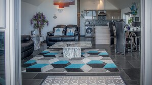 A modern living room with a sofa, a coffee table, and a rug. There is a kitchen in the background with a washing machine and a dining area with chairs. - Nautilus - 2 Bedroom Apartment - Pendine (Carmarthen)