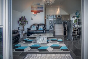 A modern living room with a sofa, a coffee table, and a rug. There is a kitchen in the background with a washing machine and a dining area with chairs.