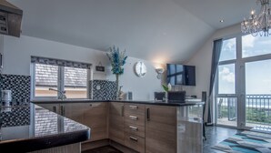 A modern kitchen with a large island, wooden cabinets, a tiled backsplash, and a view of the outdoors through large windows. - Nautilus - 2 Bedroom Apartment - Pendine (Carmarthen)
