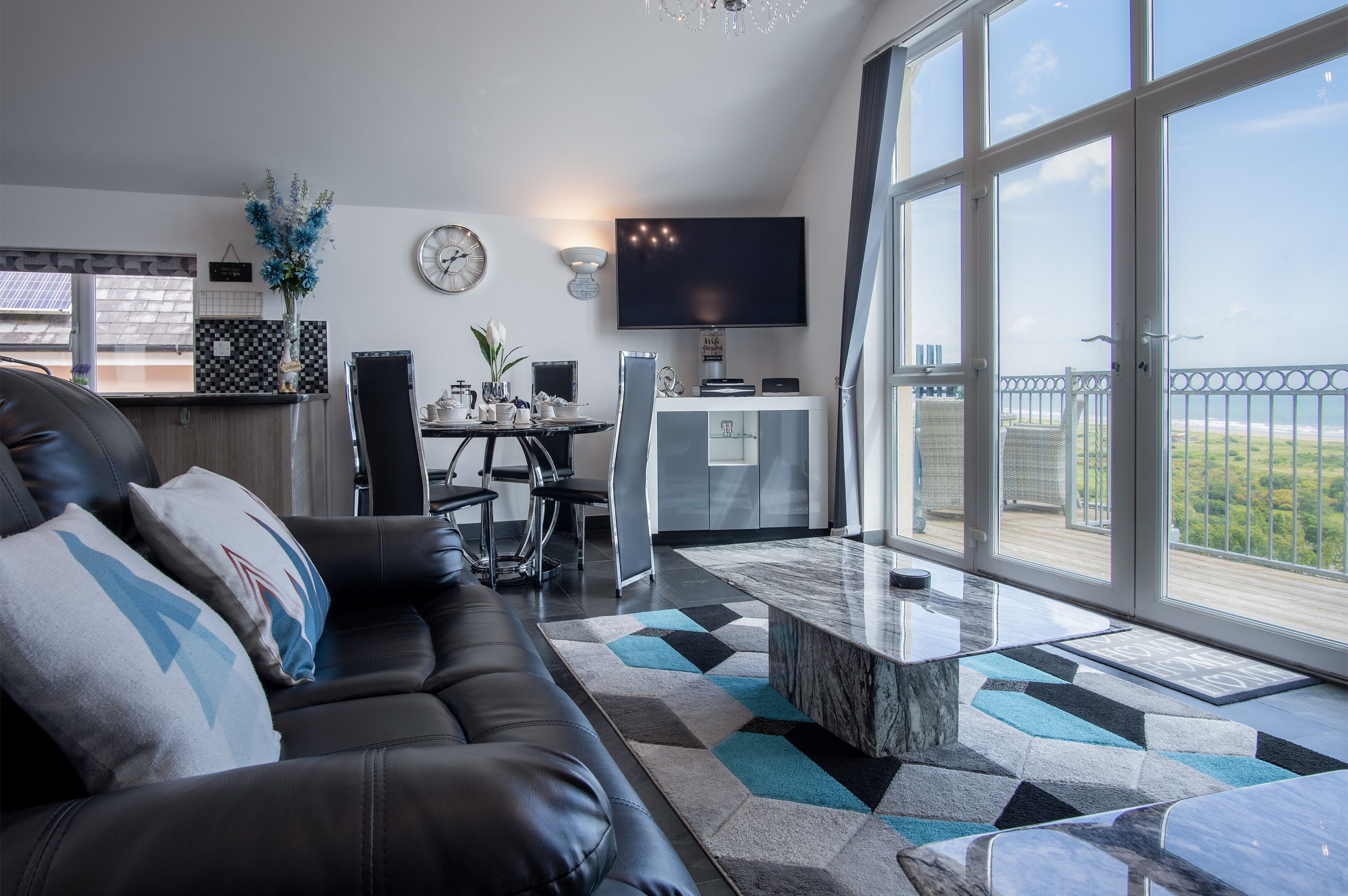 A modern living room with a black leather sofa, a dining area with a table and chairs, a flat-screen TV mounted on the wall, and large windows offering a view of the outdoors.
