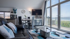 A modern living room with a black leather sofa, a dining area with a table and chairs, a flat-screen TV mounted on the wall, and large windows offering a view of the outdoors. - Nautilus - 2 Bedroom Apartment - Pendine (Carmarthen)