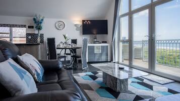 A modern living room with a black leather sofa, a dining area with a table and chairs, a flat-screen TV mounted on the wall, and large windows offering a view of the outdoors.