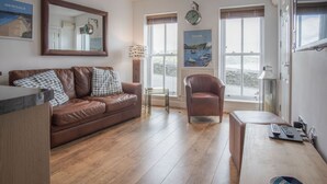 A living room with a brown leather sofa, a glass coffee table, a brown armchair, and a wooden bench. There are two windows with blinds, a wall clock, and a poster of a coastal scene.