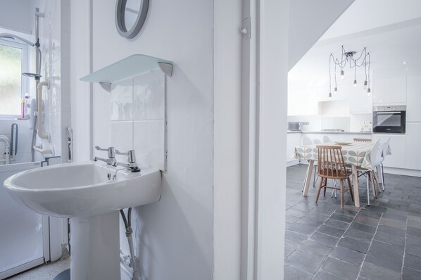 A modern bathroom with a white sink and a glass shelf, a shower area, and a view into a dining area with a table and chairs. - Ty Golau - 3 Bed Holiday Home - Pembroke (Pembroke)