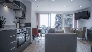 A modern kitchen with built-in appliances, a dining area with wicker furniture, and a large window with a view of the outdoors. - Beach Court - 1 Bedroom Apartment - Saundersfoot (Saundersfoot)