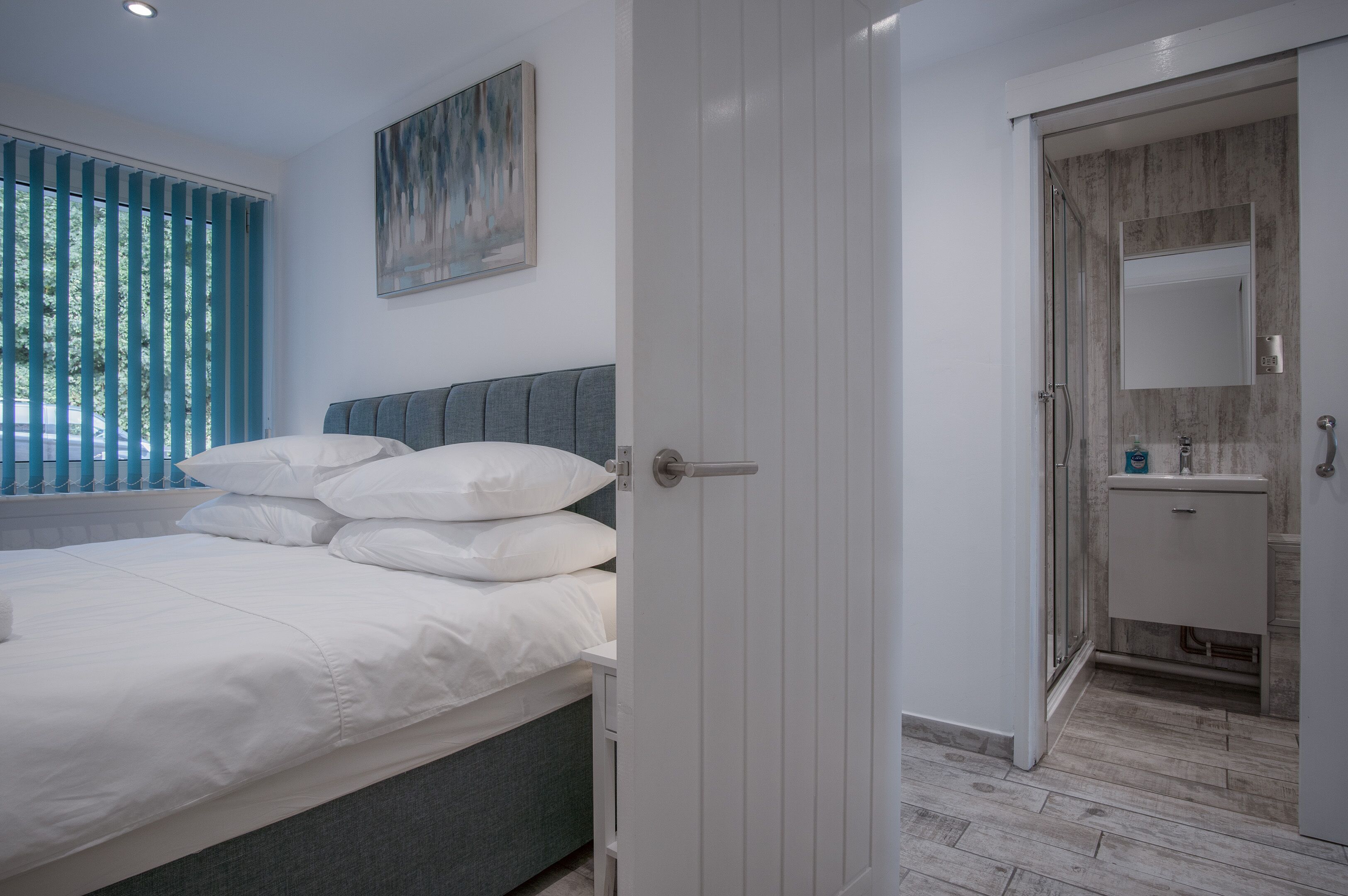 A bedroom with a bed, a gray headboard, a white door, and a bathroom with a sink and mirror.