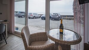 A room with a large glass door leading to a parking area and a view of the sea. There is a wicker chair and a table with a bottle and glass. - Beach Court - 1 Bedroom Apartment - Saundersfoot (Saundersfoot)
