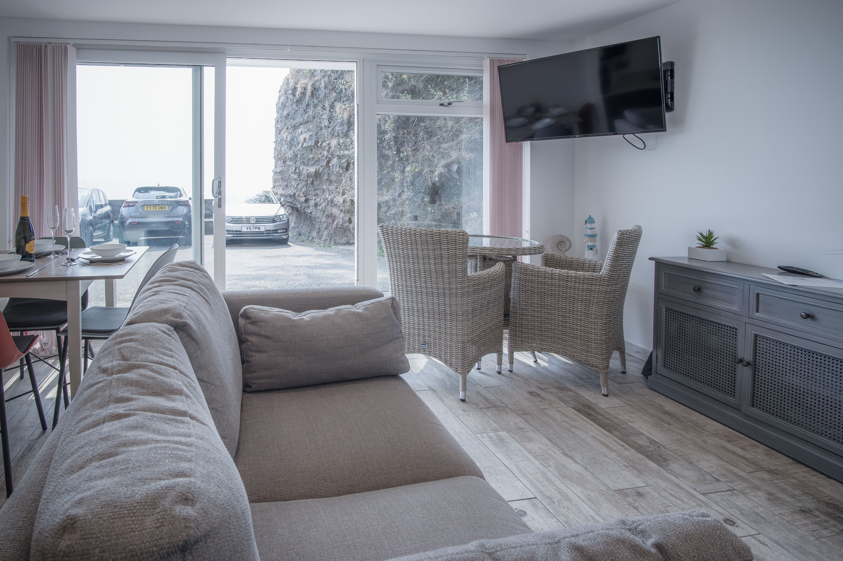 Ein modernes Wohnzimmer mit einer Couch, Korbstühlen, einem Esstisch, einem an der Wand montierten Fernseher und Blick auf geparkte Autos und einen Berg.