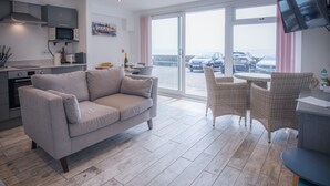 A modern living room with a sofa, wicker chairs, a dining table, and a kitchen area with a microwave and cabinets. - Beach Court - 1 Bedroom Apartment - Saundersfoot (Saundersfoot)