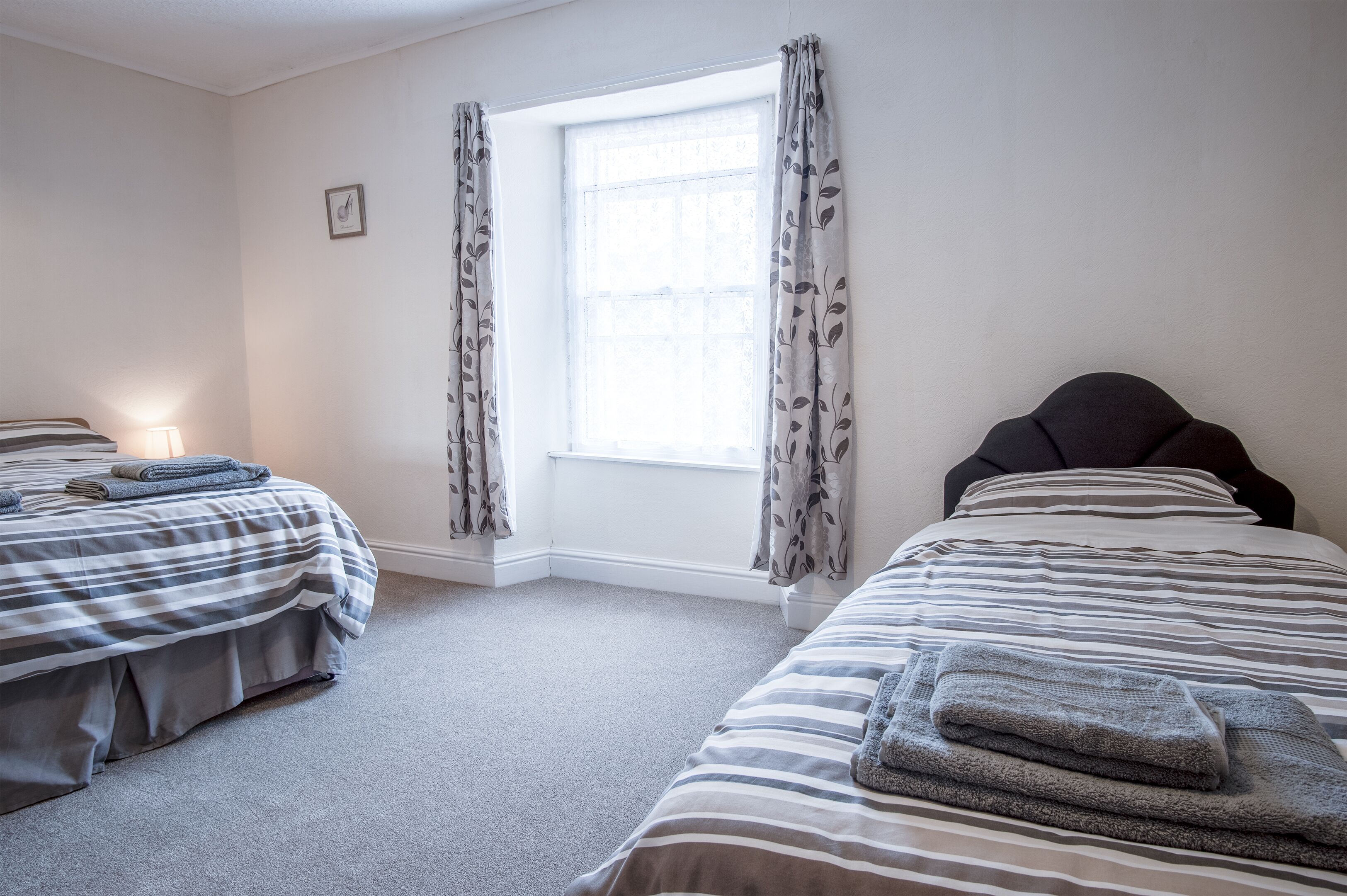 A bedroom with two beds, a window with curtains, a bedside lamp, and a framed picture on the wall.