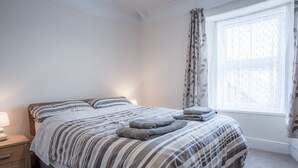A neatly made bed with striped bedding, a bedside lamp, and a window with patterned curtains. - Dragons Den - 2 Bedroom Apartment - Tenby (Tenby)