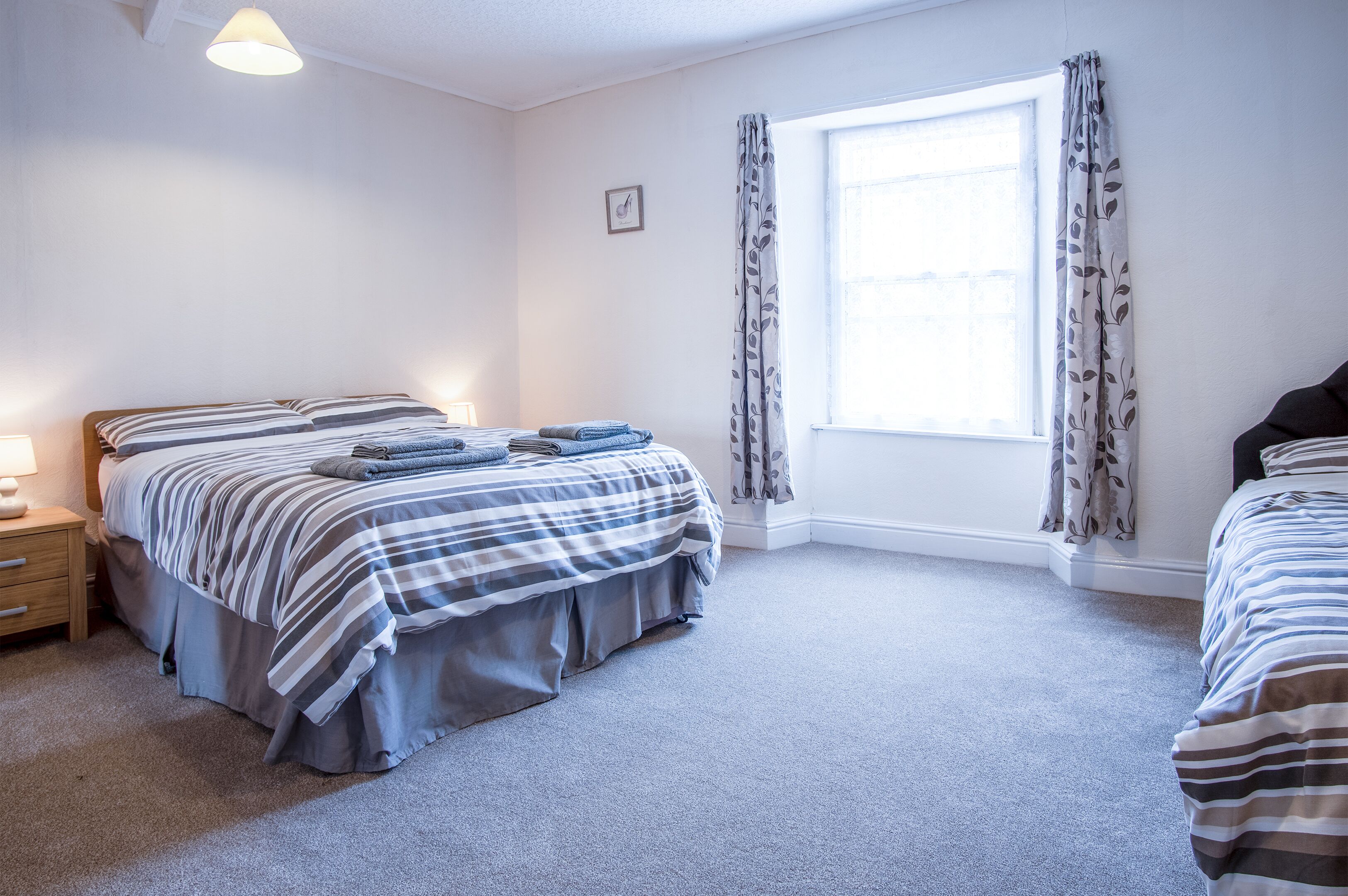 A bedroom with a bed, a bedside table, a window with curtains, and a carpeted floor.