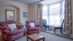 A living room with two armchairs, a small table, and a window with curtains. - Dragons Den - 2 Bedroom Apartment - Tenby (Tenby)