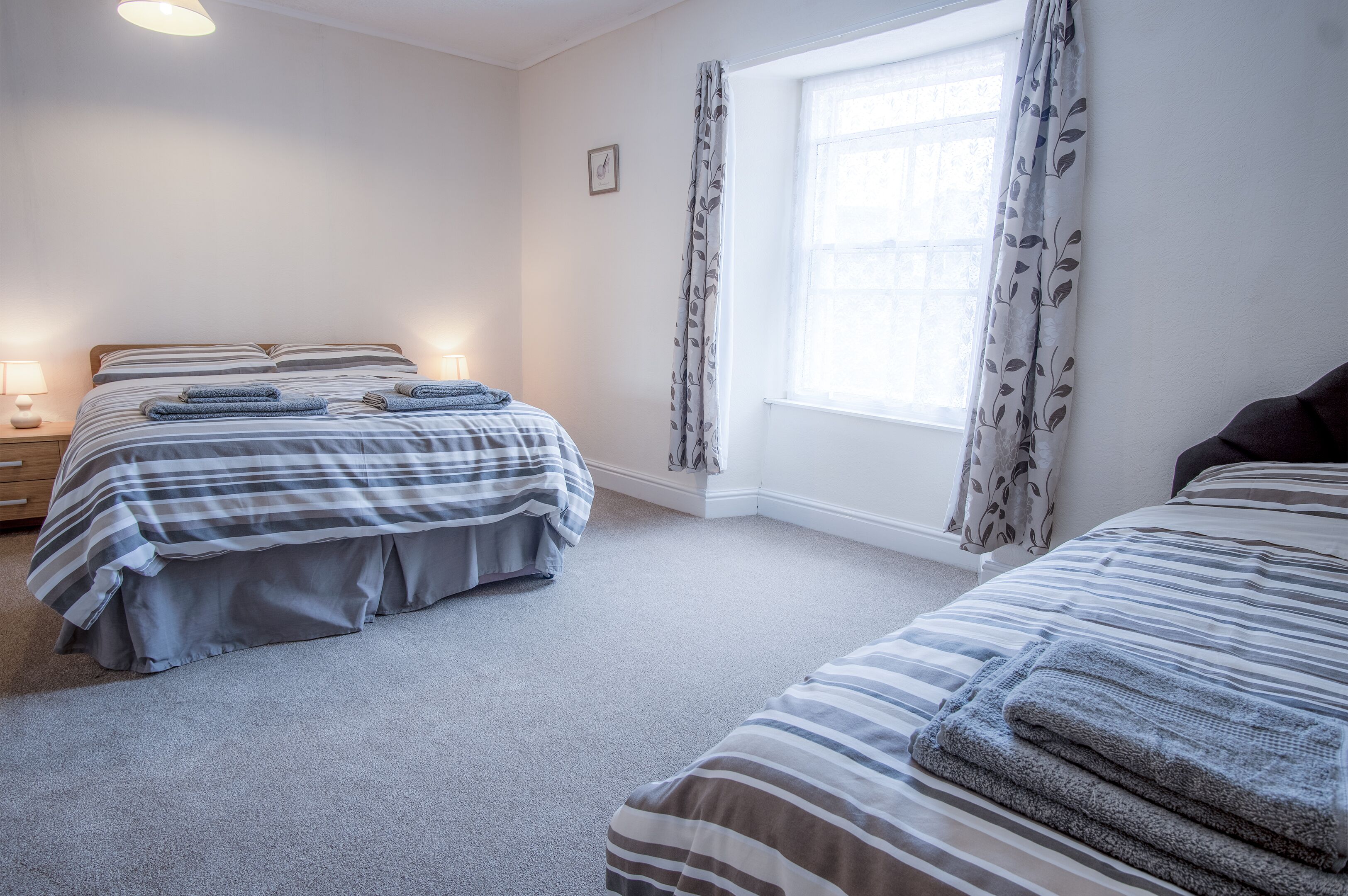 A bedroom with two beds, a window with curtains, a bedside table with a lamp, and a small picture on the wall.