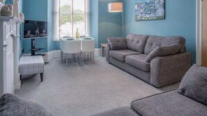 Ein Wohnzimmer mit grauem Sofa, einem kleinen Tisch und einer Bank. An der Wand hängt ein Gemälde und es steht eine Lampe dort.