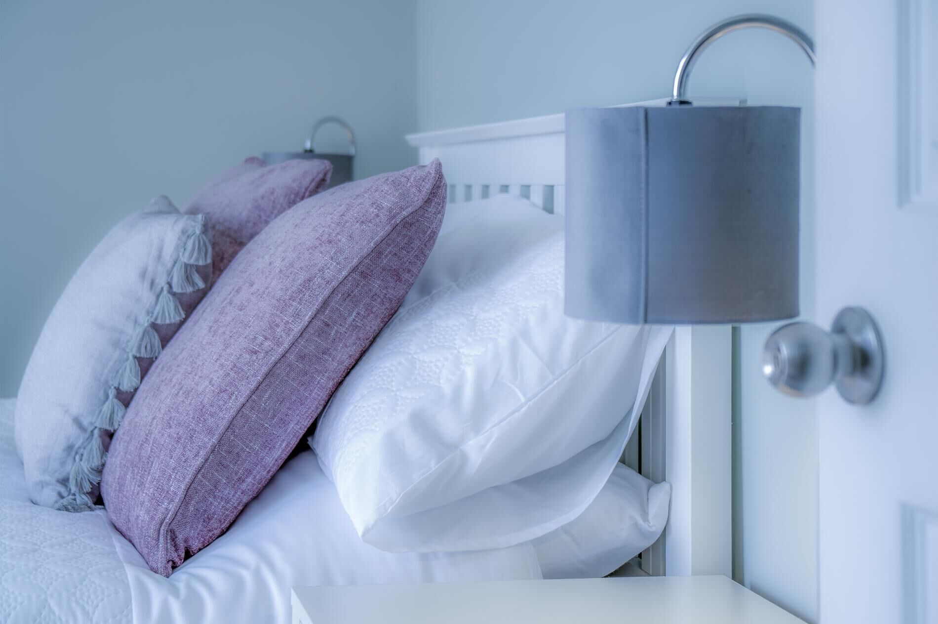 A neatly made bed with white and purple pillows, a grey bedside lamp, and a white nightstand.