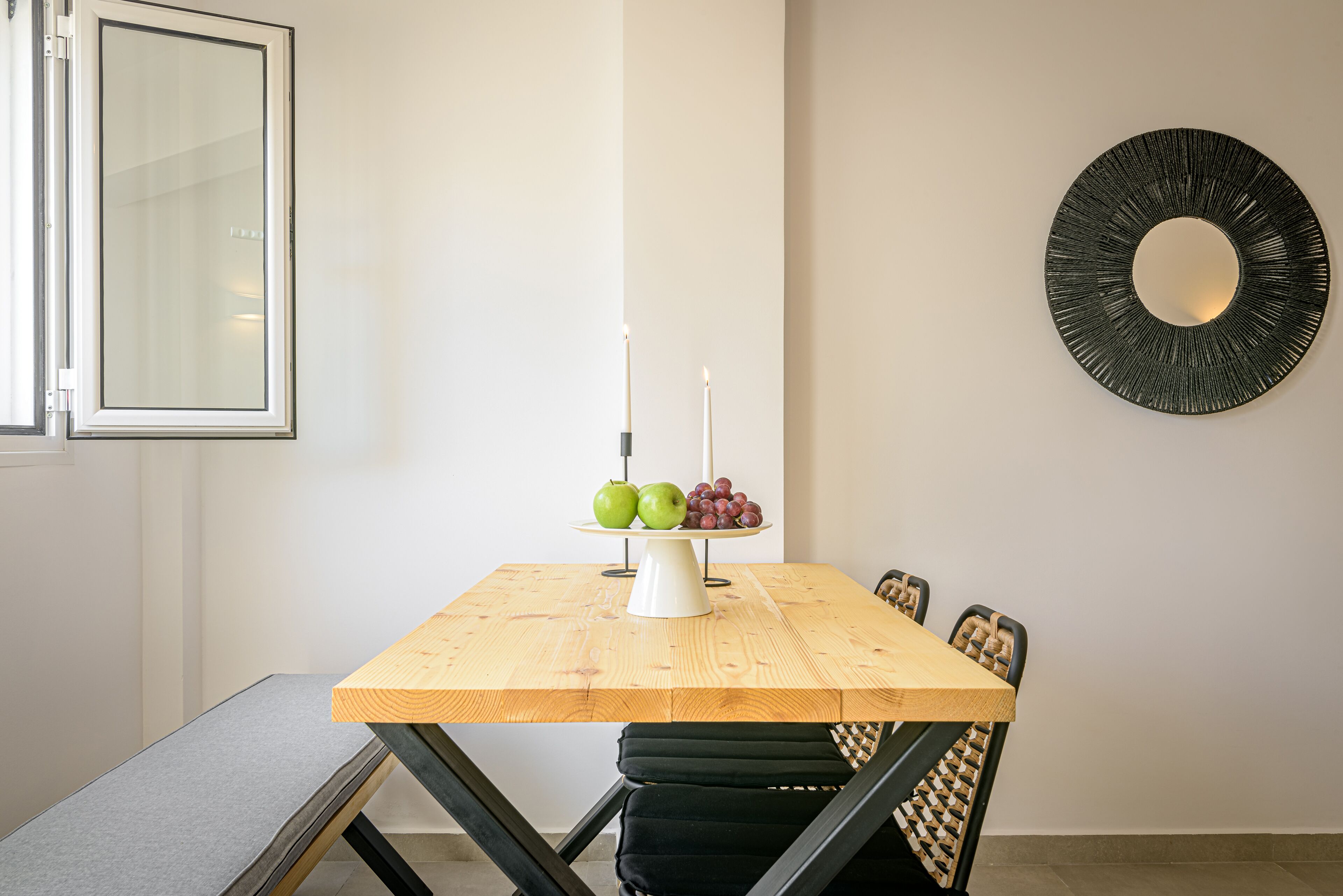A dining area with a wooden table, black chairs, a white candle holder with fruit, and a round mirror on the wall.