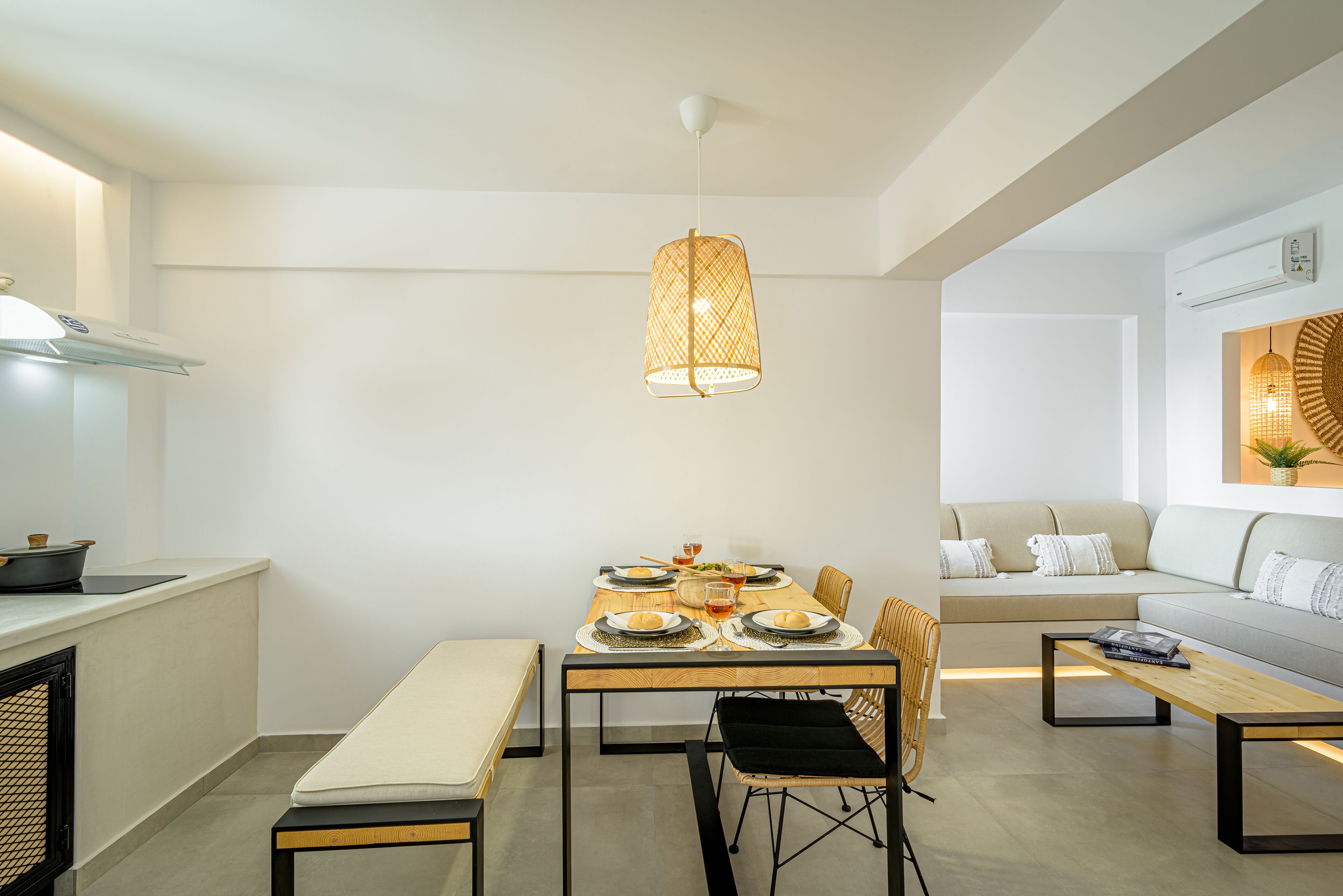 A modern dining area with a wooden table set for four, surrounded by chairs. A pendant light hangs above the table, and a sofa is visible in the background.