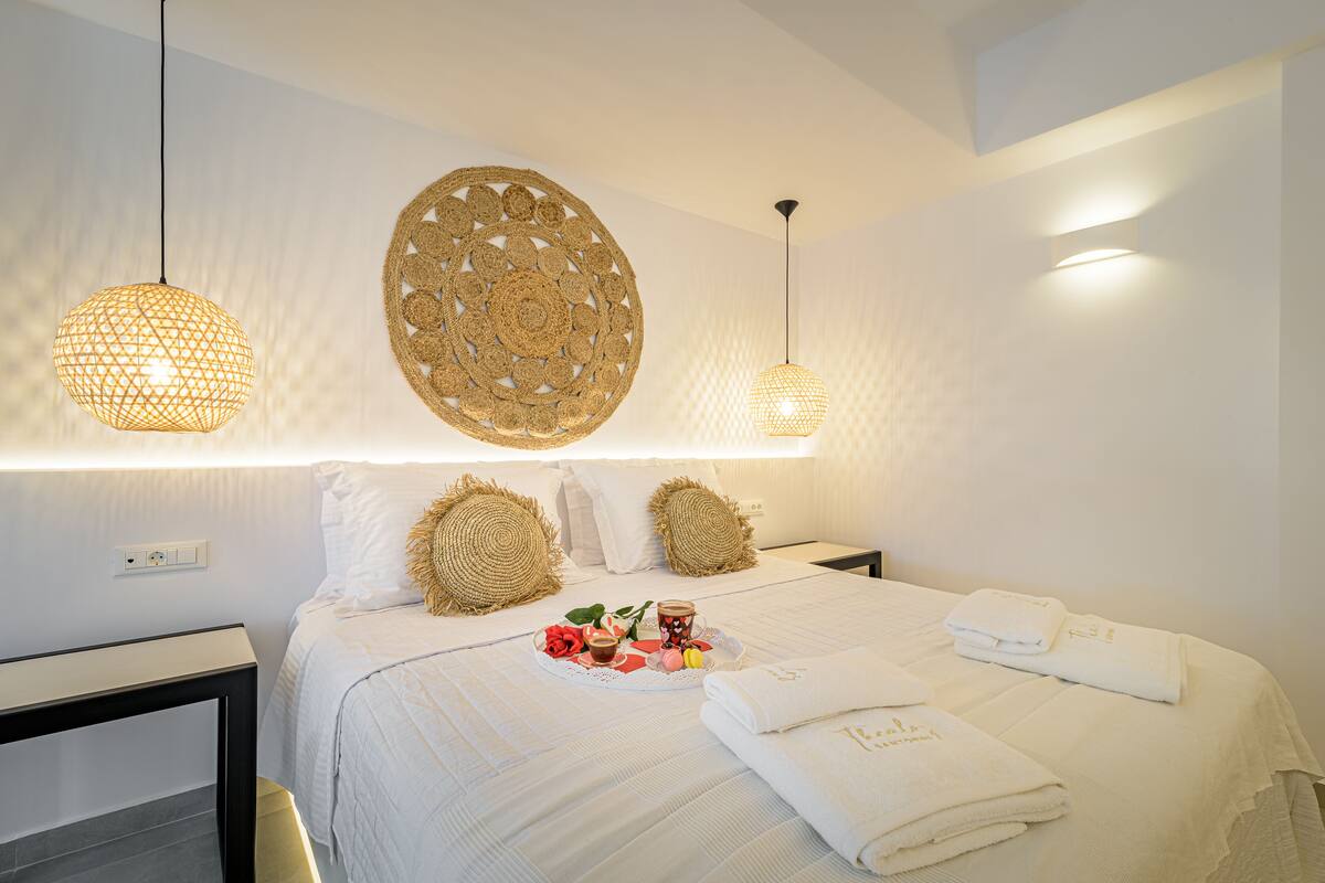 Uma cama bem arrumada com roupa de cama branca, uma mesinha de cabeceira e um objeto decorativo fixado na parede.