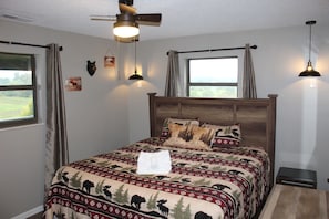 A bedroom with a large bed, a ceiling fan, two windows with curtains, and wall-mounted lights.