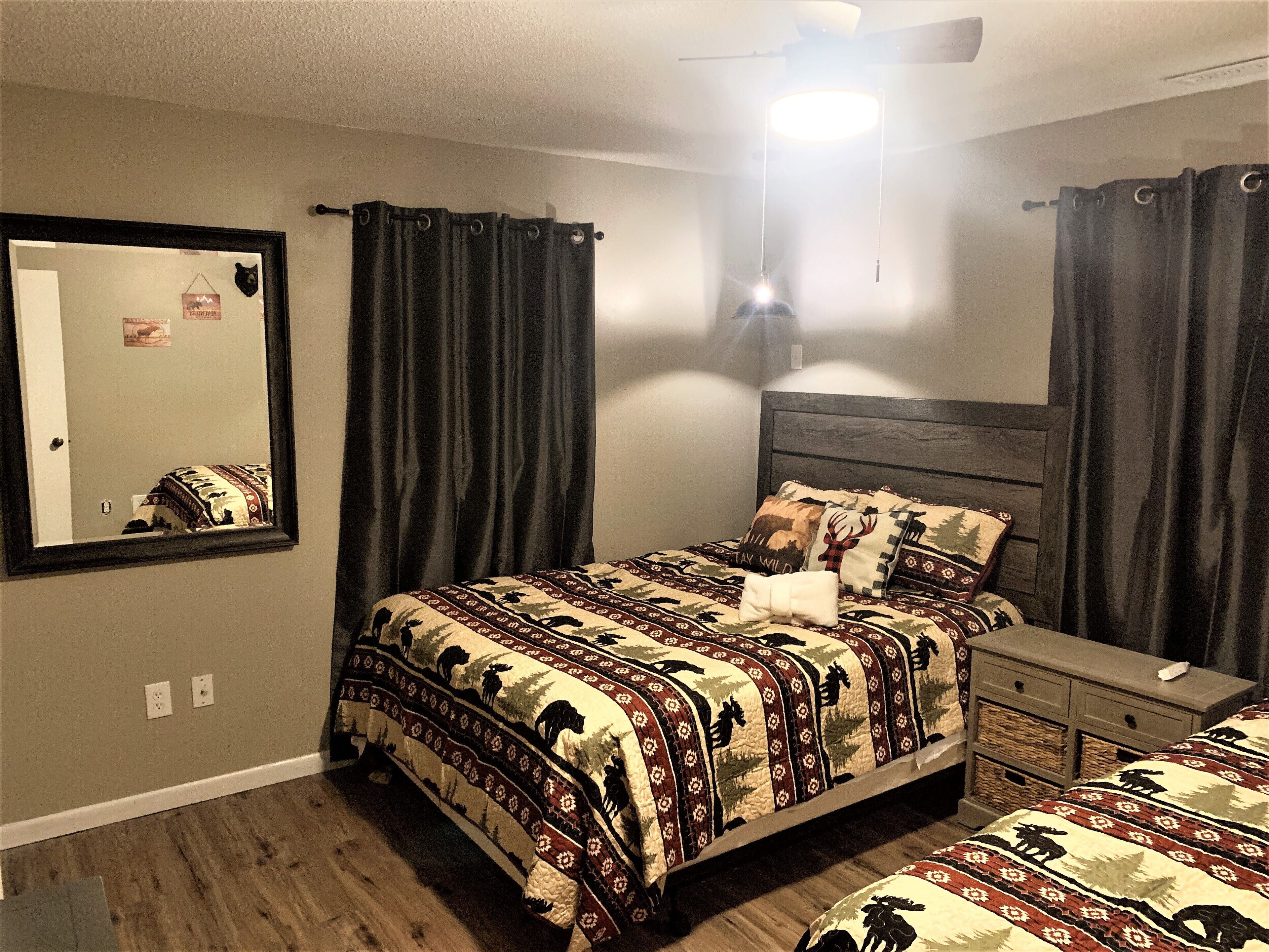 A bedroom with a large bed, a nightstand, a dresser, a mirror, and dark curtains.