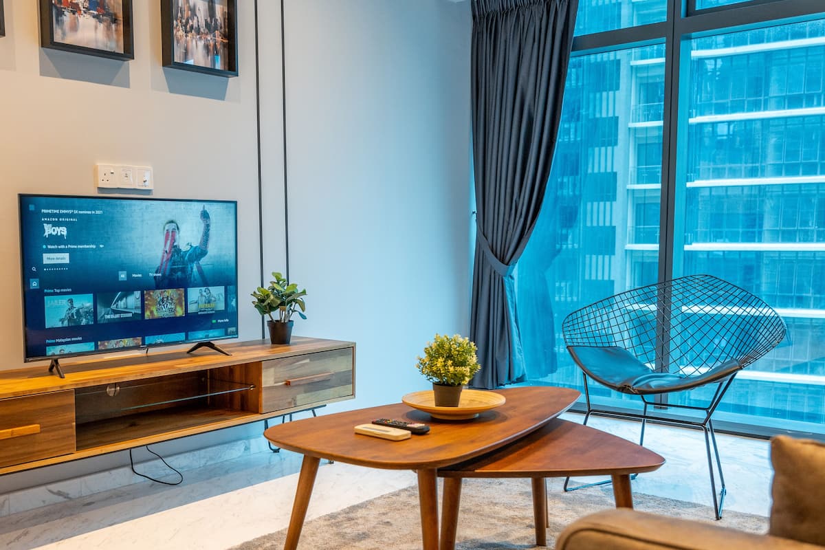 A modern living room with a flat-screen TV, wooden furniture, a potted plant, and a view of a cityscape through large windows.