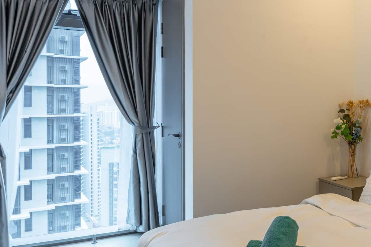 A hotel room with a bed, a nightstand, a vase with flowers, and a view of tall buildings through the window.