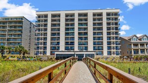 A multi-story hotel with a central entrance, a wooden walkway leading to the entrance, and a grassy area with palm trees. - Welcome to WATER POINTE II, your ideal oceanfront escape in North Myrtle Beach (North Myrtle Beach)