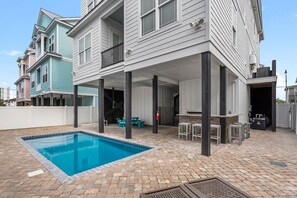 Una casa de dos pisos con un patio cubierto, una piscina y mobiliario exterior.