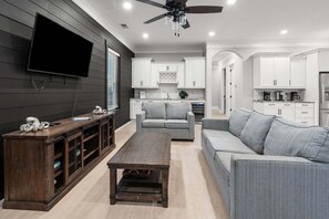 A modern living room with a flat-screen TV, grey sofas, a wooden coffee table, and a ceiling fan.