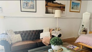 Una sala de estar con un sofá marrón oscuro, una mesa de centro de vidrio con revistas, una lámpara blanca y obras de arte enmarcadas en la pared.