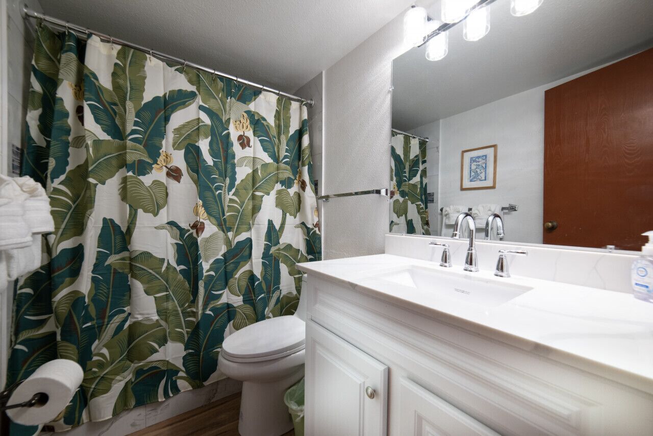 A bathroom with a shower curtain featuring a tropical leaf pattern, a white toilet, a white sink with a mirror above it, and a wooden door.