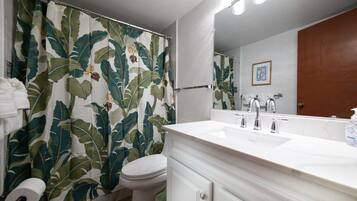 A bathroom with a shower curtain featuring a tropical leaf pattern, a white toilet, a white sink with a mirror above it, and a wooden door.
