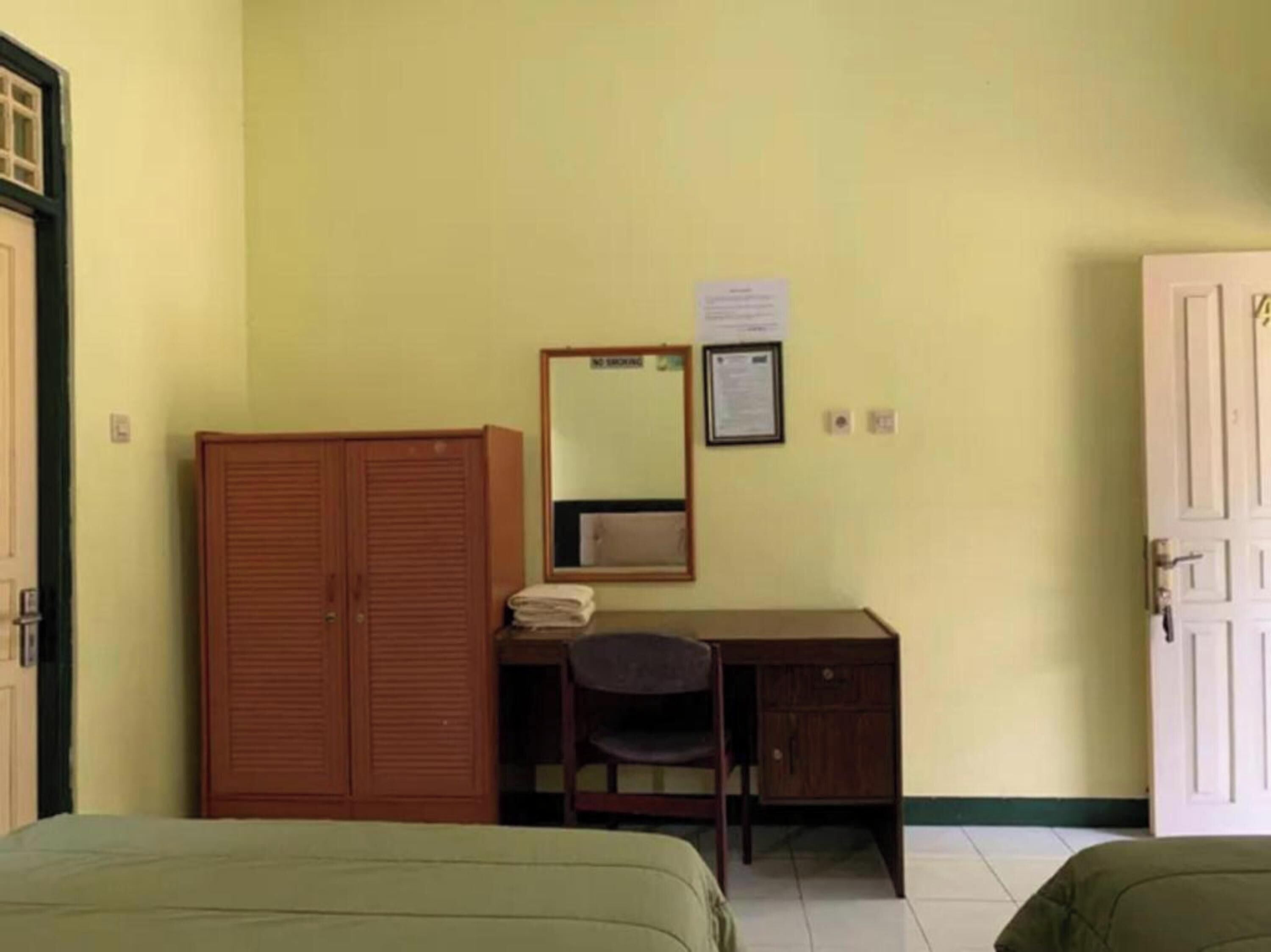 A room with a wooden wardrobe, a desk with a chair, a bed, and a door.