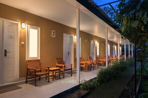 A covered outdoor seating area with wooden chairs and tables, white doors, and a garden path. - Rinjani Garden Syariah by ecommerceloka (Sembalun Lawang)