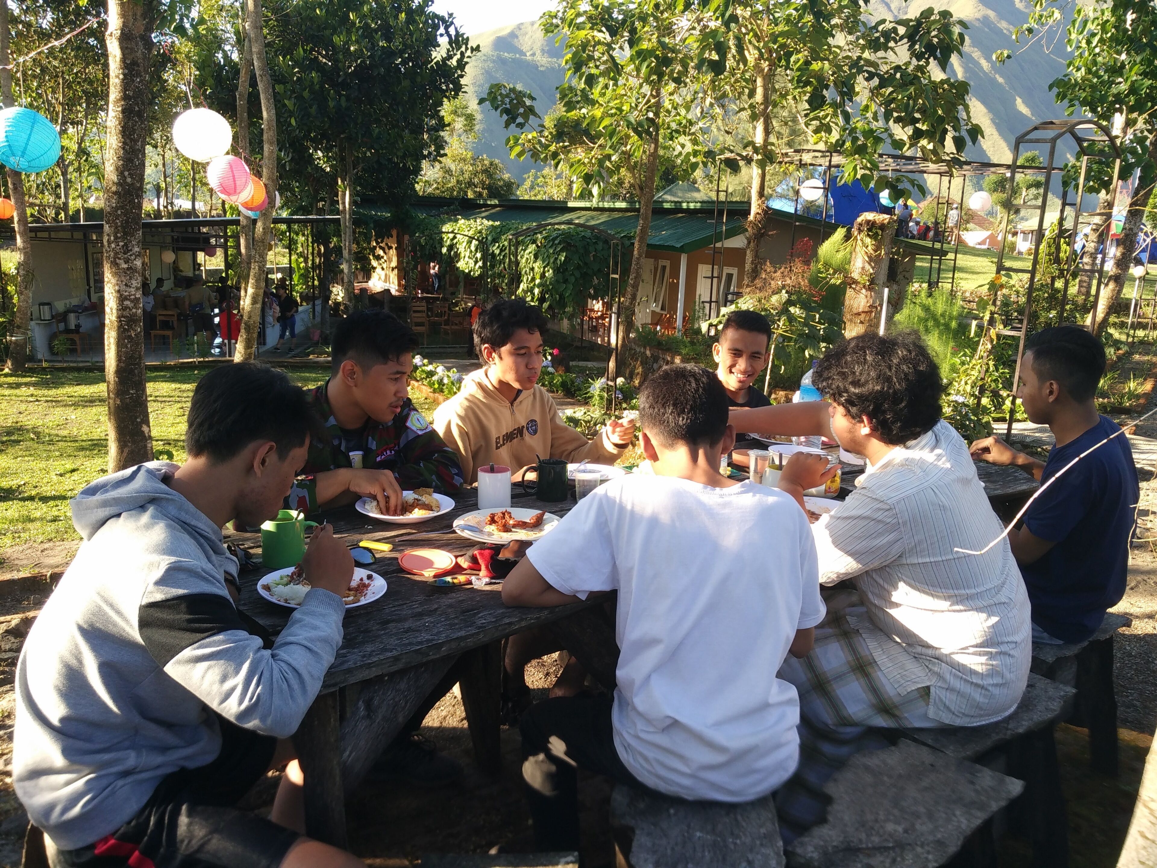 Un grupo de personas cenando al aire libre en una mesa de madera, con una montaña al fondo.
