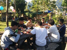 Un grupo de personas cenando al aire libre en una mesa de madera, con una montaña al fondo.