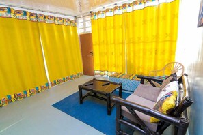 A room with yellow curtains, a sofa, a small table, and a patterned rug. - Stay.Plus Shyrah Glass Cottages (Naivasha)