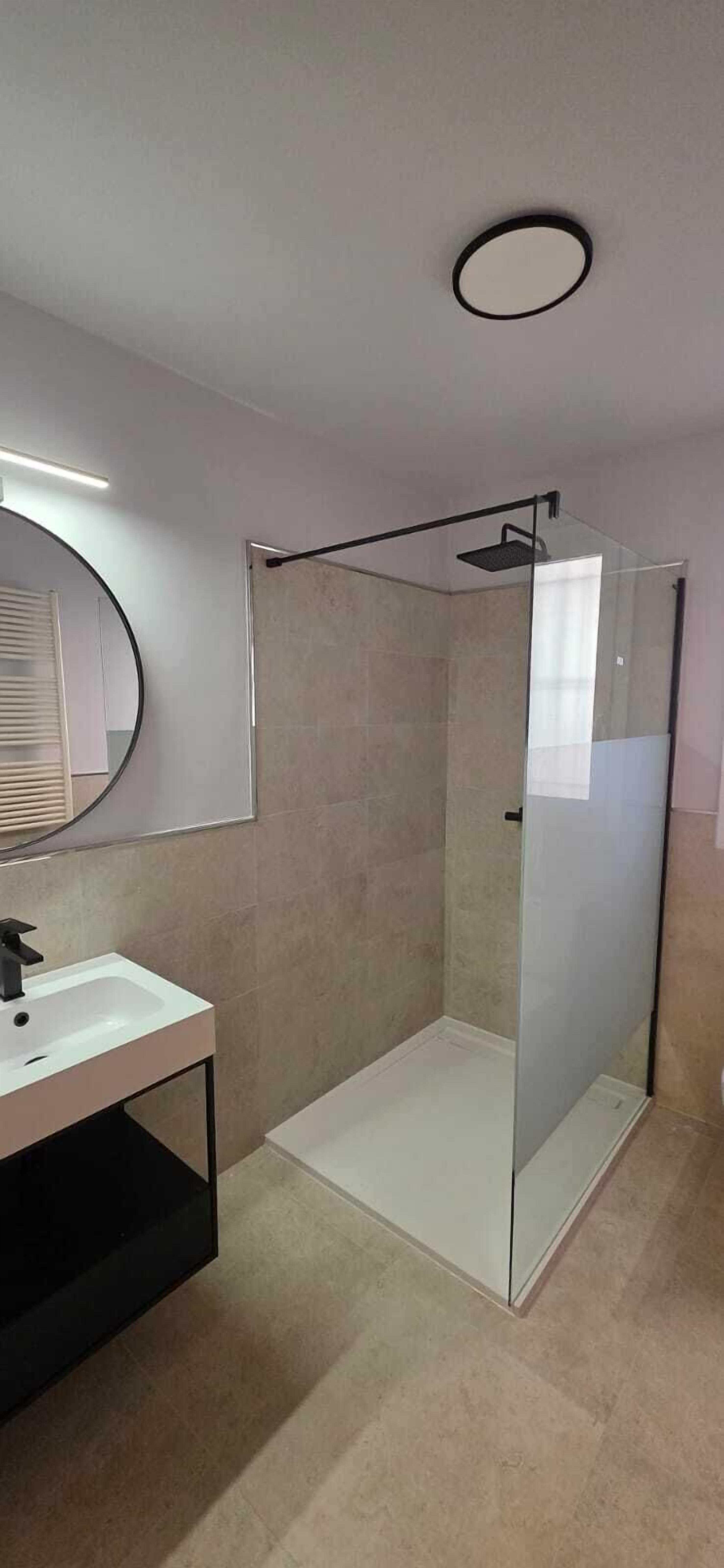 A modern bathroom with a glass-enclosed shower, a round mirror, and a white sink.