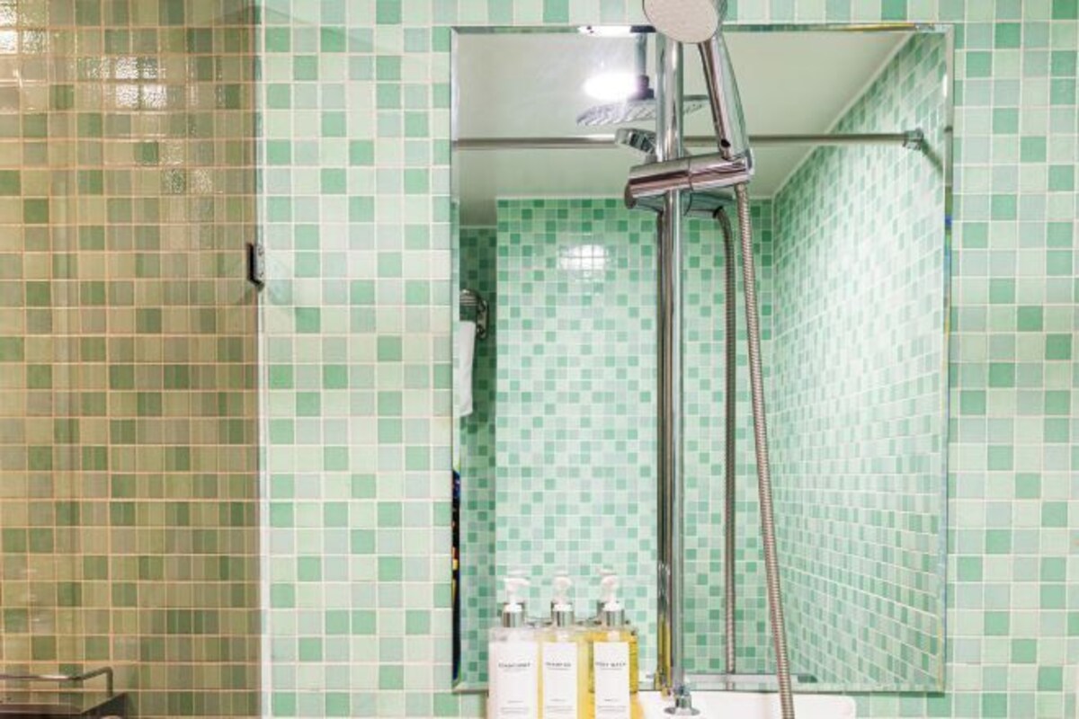 Un baño con baldosas verdes a cuadros, una ducha con cabezal de ducha manual y artículos de aseo en el lavamanos.