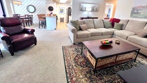A spacious living room with a large sofa, a dark leather armchair, and a coffee table. There is a kitchen area in the background with a dining table and chairs.