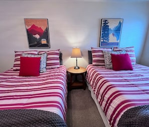 A room with two beds, each with striped bedding and decorative pillows, a small round table with a lamp, and two wall-mounted posters. - Park Place Aspen Condo • Walk to Main St. • Snowflake Lift • Pool+Hot Tubs (Breckenridge)
