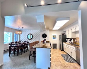 A modern kitchen with white cabinets, a stainless steel refrigerator, and a dining area with a wooden table and chairs. - Park Place Aspen Condo • Walk to Main St. • Snowflake Lift • Pool+Hot Tubs (Breckenridge)