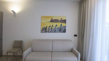 Un salon avec un canapé beige, un fauteuil, un tableau représentant un paysage côtier et une fenêtre avec des rideaux.