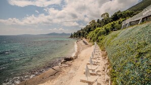 Una playa costera con un paseo marítimo pavimentado, sillas de playa y un muro de piedra con vegetación.