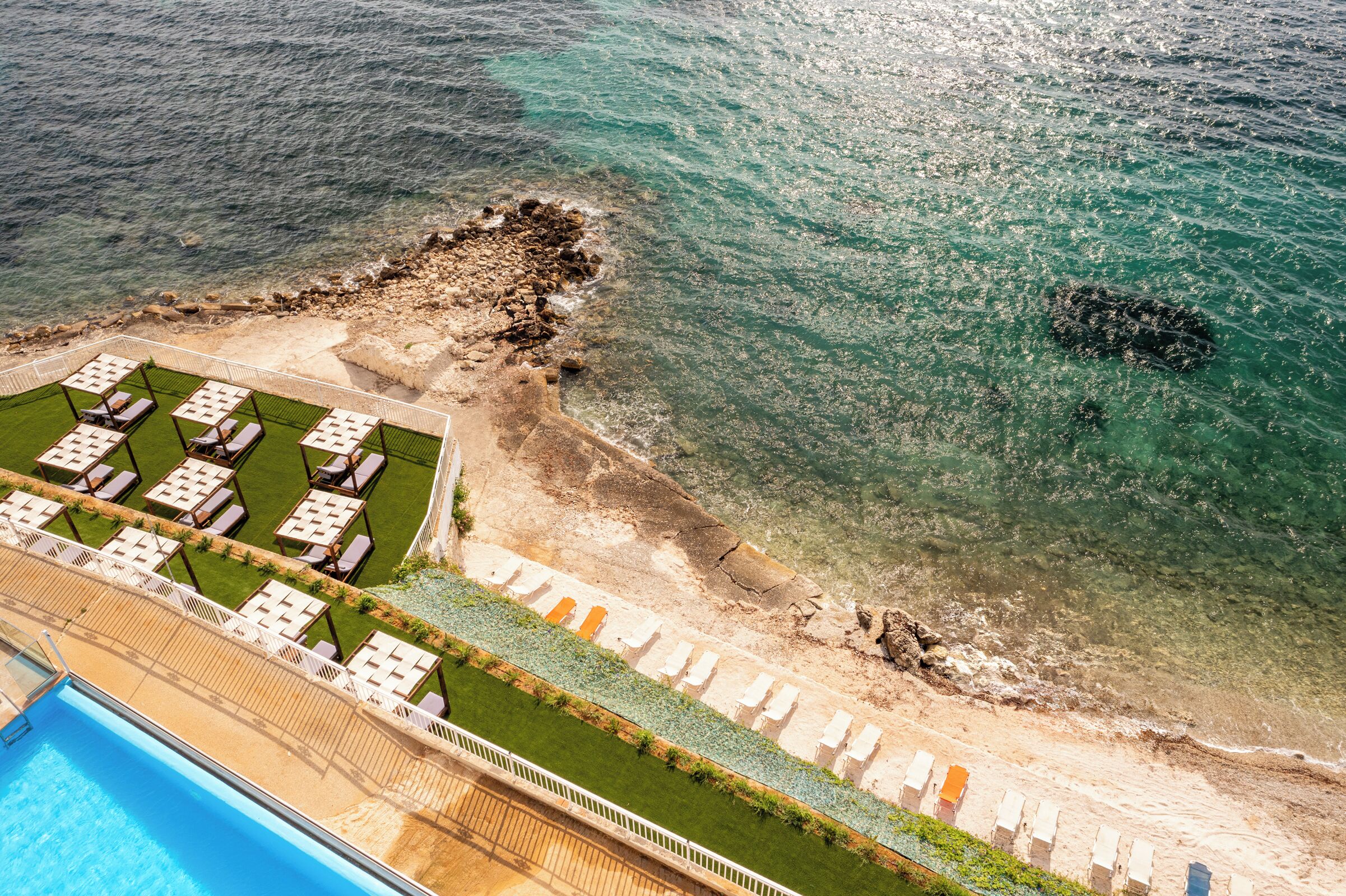 Vista aerea di un resort con piscina, lettini prendisole e spiaggia.