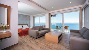 Una sala de estar moderna con un sofá, una mesa de centro y una cama con una colcha roja. Hay un ventanal con vistas al mar.