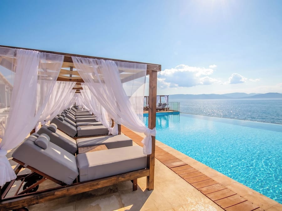A poolside lounge with a view of the sea, a wooden deck, and a canopy with white fabric.
